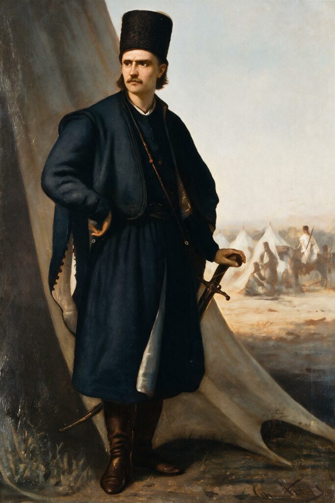 Full-length painted portrait of Tudor Vladimirescu by Theodor Aman. He stands beside a large canvas tent, wearing a dark blue Wallachian coat and a tall black fur hat, one hand resting on the hilt of a sabre, his gaze turned to the side. Behind him a military camp of white tents and small figures recedes into the distance.